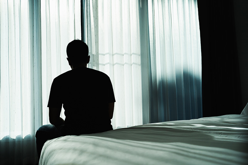A man struggling with mental health stigma for men sits at the edge of his bed and faces away from the camera toward a window.